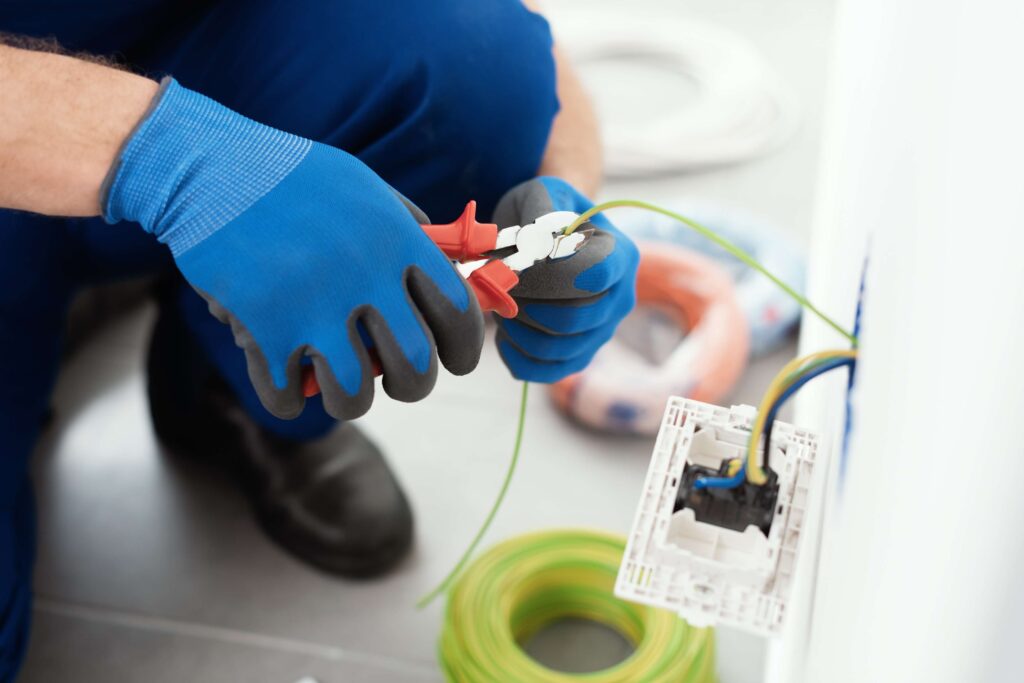 Electrician repairing house wiring with insulated gloves and wire cutters near an open wall outlet.