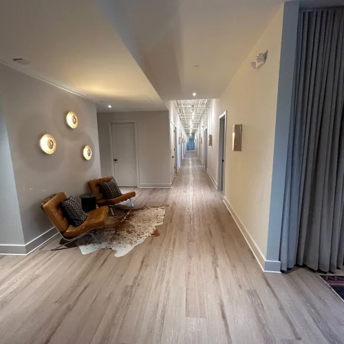 A modern hallway featuring light wood flooring, decorative wall lights, and two brown chairs with decorative pillows beside a rug.