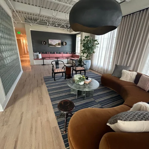 A modern lounge features a curved brown sofa, striped rug, coffee table, and stylish chairs, with large windows and plants.