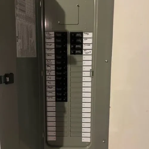 An electrical panel with a gray metal door open, displaying several labeled circuit breakers and a wiring diagram on the left.