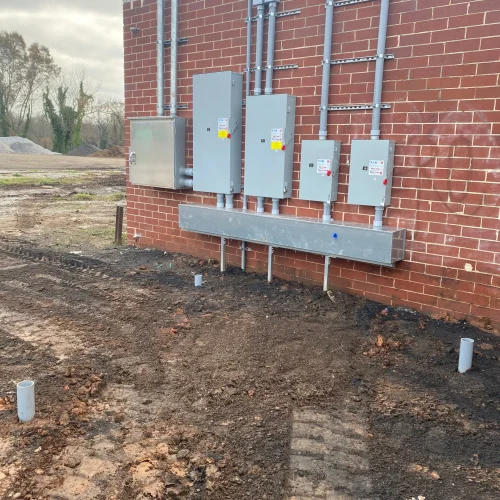 Electrical panels and conduits attached to a red brick wall, with a cleared ground area and a few concrete pipes visible.
