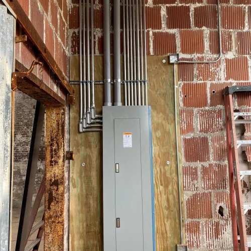 A gray electrical panel mounted on a wooden board, with multiple metal conduits running vertically along a brick wall.
