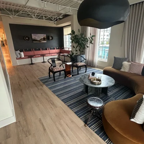 A modern lounge features a curved brown sofa, striped rug, coffee table, and stylish chairs, with large windows and plants.