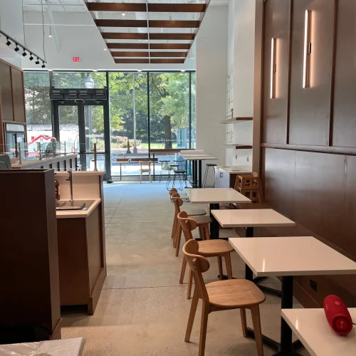 Modern cafe interior featuring wooden chairs and white tables, with large windows letting in natural light.