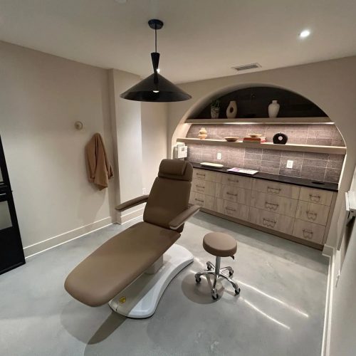 A modern treatment room featuring a reclining chair, a stool, and a sleek counter with decorative items and soft lighting.